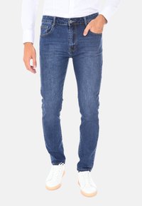Uomo che indossa jeans blu slim-fit e sneakers bianche, con una mano in tasca e una camicia bianca a maniche lunghe.
