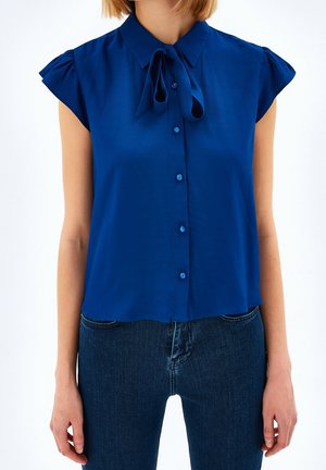Blouse bleue à manches courtes avec boutons et col noué en forme de nœud, portée sortie sur un jean en denim bleu foncé.