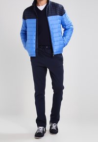 Blå puffjacka med en svart panel vid axlarna, dragkedjestängning och marinblåbyxor. Sneakers har vita accenter och snörningar.