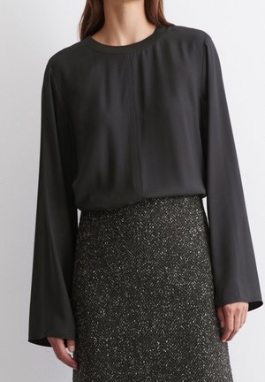 Kvinde iført sort bluse med lange ærmer og rund halsudskæring samt tekstureret mørk tweed nederdel.