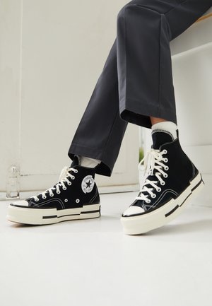 Baskets montantes Converse noires avec lacets et semelles blanches portées avec des chaussettes blanches et un pantalon gris foncé à l'intérieur.