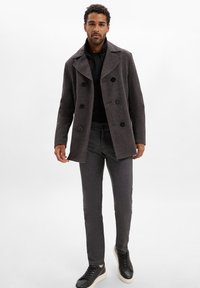 Man in een donkergrijze dubbelzijdige jas, zwarte shirt, grijze slim-fit broek en zwarte sneakers met witte zolen, staand tegen een witte achtergrond.
