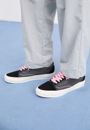 Black and gray sneakers with pink laces and white rubber soles, featuring contrasting stitching and a low-top design, worn with gray pants.