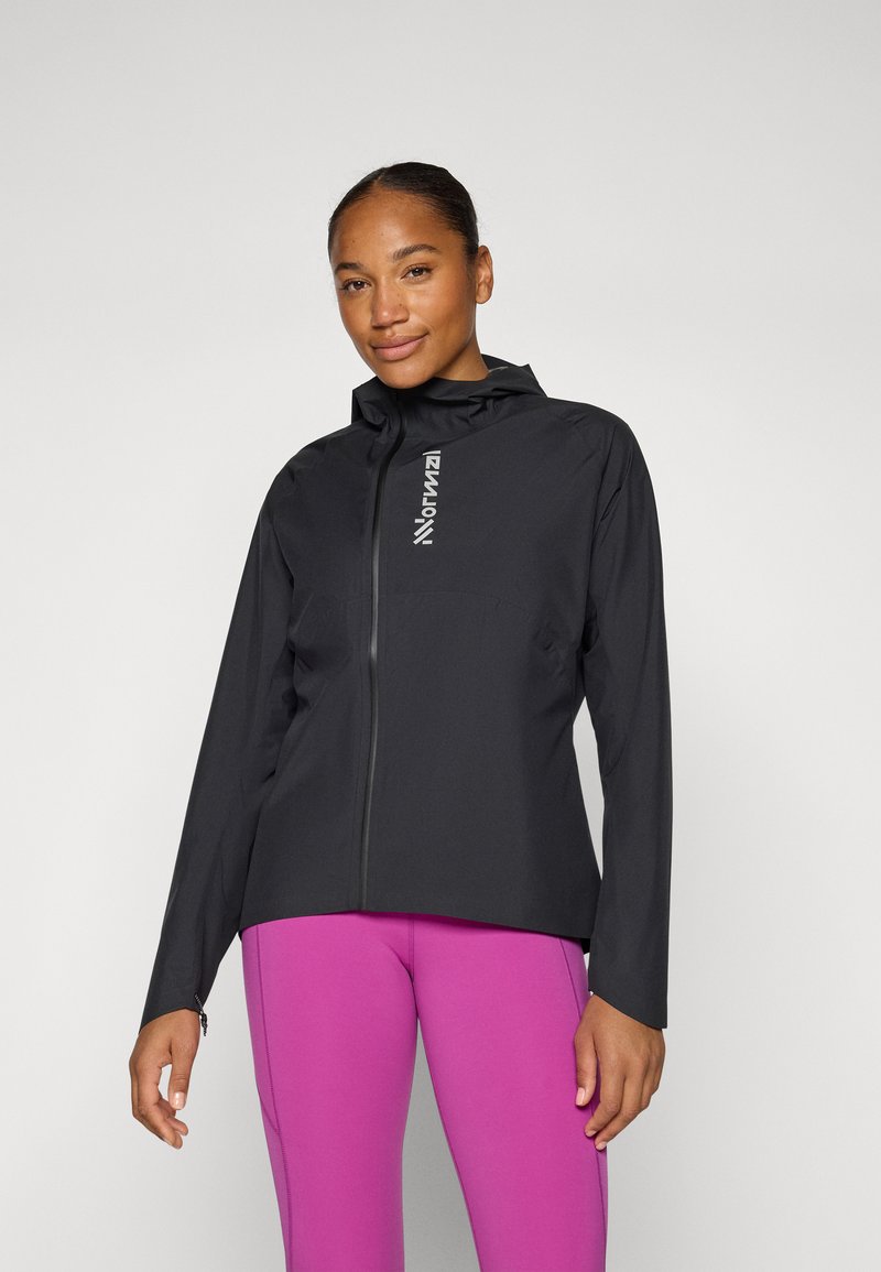 Schwarze leichte Jacke mit Kapuze, ausgestattet mit einem Reißverschluss vorne und Logodetail. Getragen über rosa Sportlegging. Glatte Textur.
