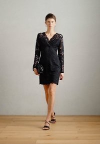 Crna čipkasta haljina s dugim rukavima, V-izrezom i prilagođenim siluetom. Uparen s crnim sandalama na remenčiće i malom clutch torbicom.