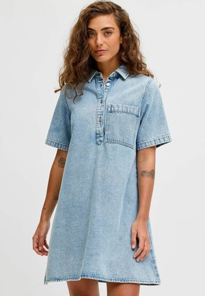 Jeune femme aux cheveux bouclés portant une robe en jean bleu clair à manches courtes avec des boutons à l'avant et une poche poitrine, debout devant un fond uni.