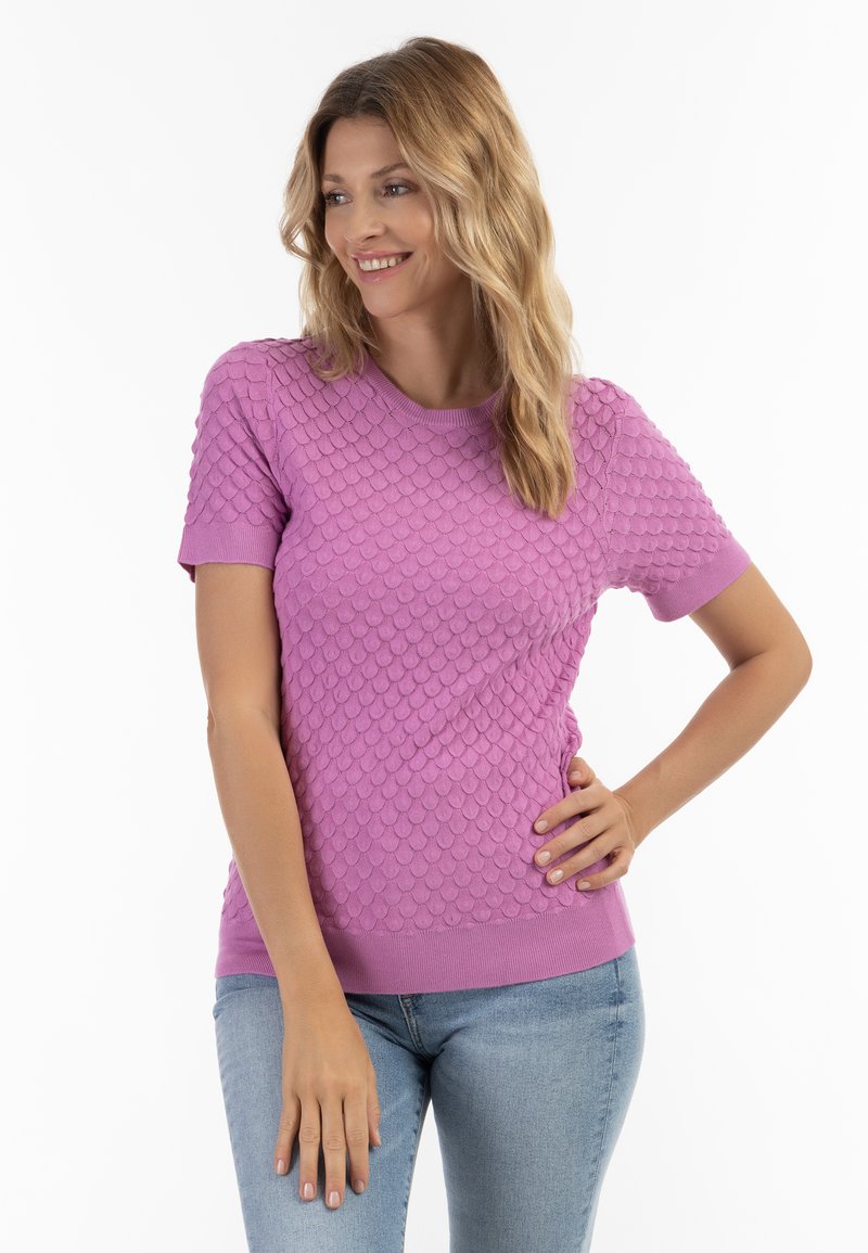 Kortærmet lilla strikket sweater med et tekstureret skælmønster, rund halsudskæring og ribbede kanter, parret med lyseblå jeans.