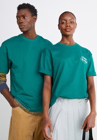Tyrkiscotton t-shirts med rulleærmer. Én har et hvidt skriftlogo. Den mandlige model bærer et flerfarvet tøj under.