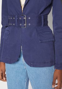 Giacca blu con cintura in tessuto resistente, dotata di ampie tasche laterali, vestibilità sartoriale e accessori in argento sulla cintura.