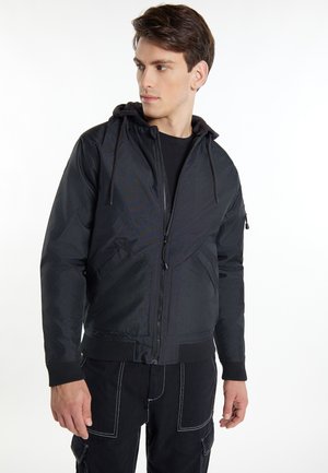 Giovane uomo con giacca nera con cappuccio e zip e pantaloni neri con cuciture bianche, guarda verso sinistra su sfondo bianco.