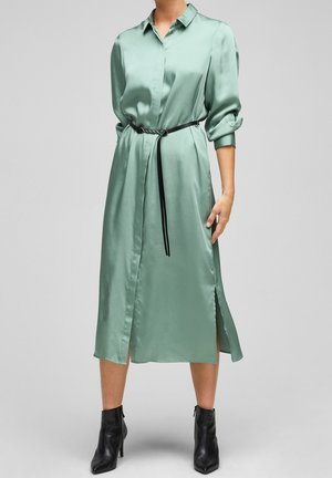 Abito camicia in satin verde menta con spacco frontale e chiusura con bottoni, stretto in vita da una cintura nera. Maniche lunghe arrotolate.