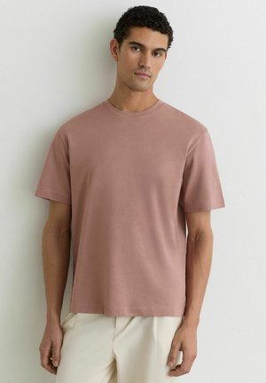 Homme aux cheveux bouclés portant un t-shirt uni rose poudré à manches courtes et un pantalon beige clair, debout contre un mur blanc.