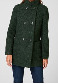 Abrigo de doble botonadura de textura verde oscuro con seis botones negros y dos bolsillos frontales, llevado sobre jeans azul oscuro.