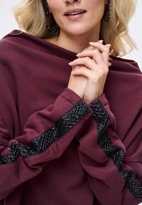 Bordeauxrode trui met lange mouwen, voorzien van gestructureerde zwarte geweven details langs de mouwen en een comfortabele, losvallende halslijn.