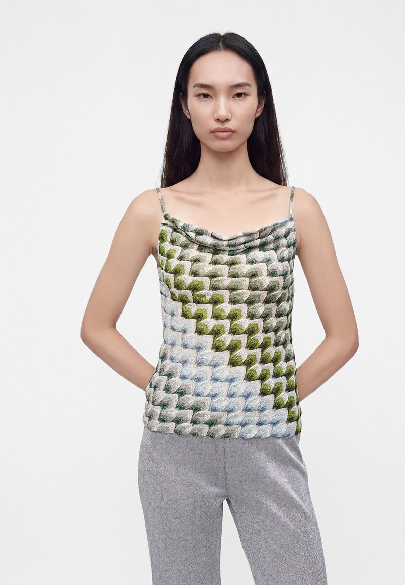 Femme aux longs cheveux noirs porte un haut sans manches à motifs avec des écailles vertes et bleues et un pantalon gris clair, debout devant un fond uni.