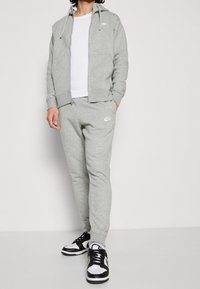 Completo tracksuit grigio Nike con felpa con zip e joggers abbinati, caratterizzato da accenti del logo bianchi. Si abbina a scarpe da ginnastica nere e bianche.
