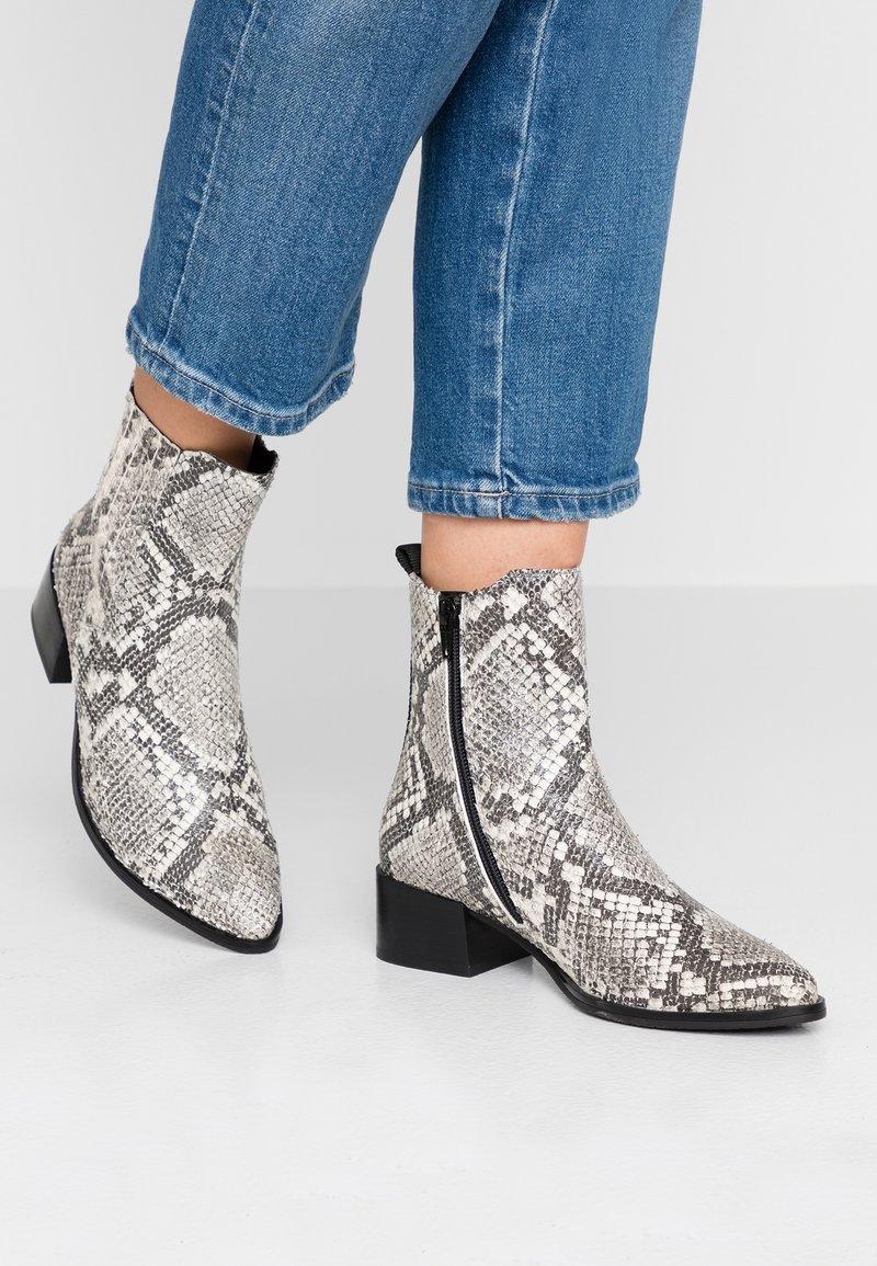 Personne portant des bottines mi-mollet à motif de peau de serpent avec des talons blocs bas et des fermetures éclair latérales, associées à un jean bleu raccourci.