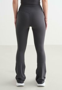 Grijze high-waisted leggings van een gladde, elastische stof met flared benen en een naaddetail aan de achterkant, gecombineerd met witte sneakers.