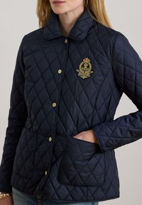 Μπλε ναυτικό καπιτονέ μπουφάν με γιακά, χρυσά μεταλλικά στοιχεία και έμβλημα. Διαθέτει δύο μπροστινές τσέπες και υφή με διαμάντι.