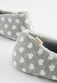 Grijze stoffen slippers met een wit hartpatroon, zachte textuur en een gestikt label met de tekst "NEXT" aan de zijkant. Driehoekige vorm met een gewatteerde rand.