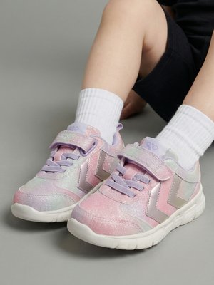Kinderbeine mit weißen Knöchelsocken und pastellrosa und violetten glitzernden Sneakers mit Klettverschluss, sitzend auf einem grauen Boden.