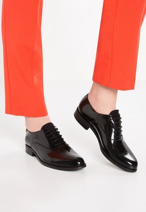 Zapatos de charol negro con cordones y textura lisa, con perforaciones decorativas en la punta, combinados con pantalones rojos brillantes.