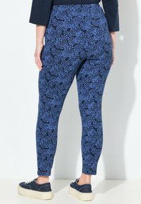 Persona che indossa leggings blu e neri con un motivo e sneakers blu scuro, in piedi contro un muro bianco, rivolta di spalle.