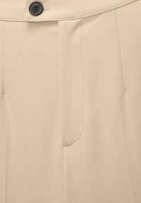 Pantalon beige sur mesure avec un bouton noir et une braguette avant cachée, montrant la texture du tissu et les détails des coutures.