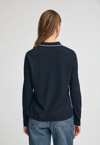 Granatowa koszula polo z długim rękawem z białymi detalami kołnierza, miękka w dotyku i luźny fason, zestawiona z jasnoniebieskimi dżinsami.