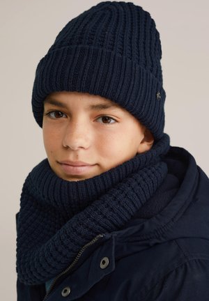Junge mit einer marineblauen Strickmütze, passendem Schal und dunkler Jacke, der leicht zur Seite schaut mit einem neutralen Ausdruck, hellen Hintergrund.