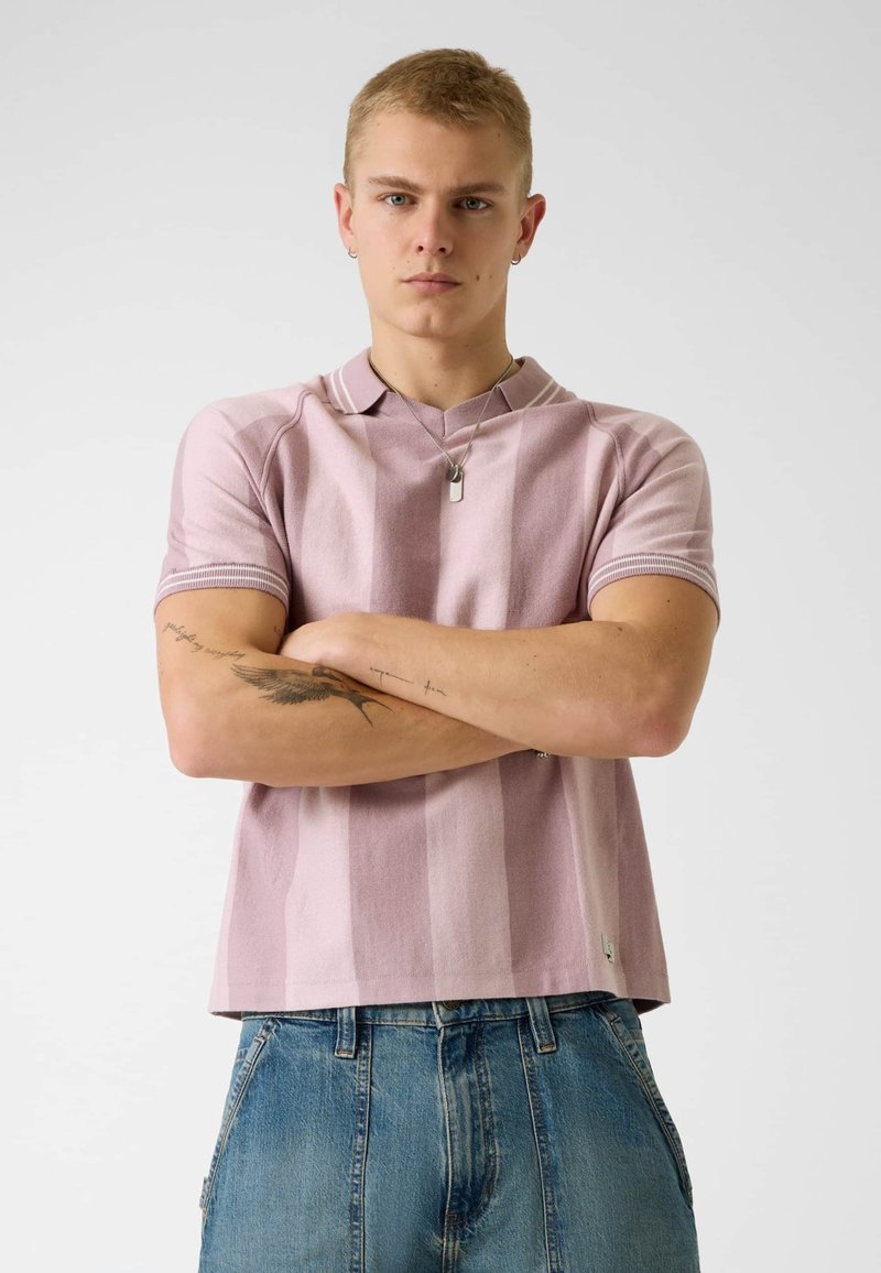 Jeune homme aux cheveux blonds courts portant un polo à rayures roses et un jean bleu, bras croisés montrant des tatouages sur les deux avant-bras.