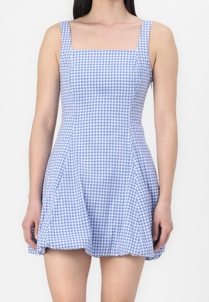 Kobieta ubrana w sukienkę bez rękawów o niebiesko-białej kratce w drobną kratkę, z kwadratowym dekoltem i rozkloszowanym dołem.