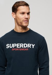 Marineblå sweatshirt lavet af en blanding af bomuld, med rund hals. Stort hvidt "SUPERDRY"-logo og røde "SPORTSWEAR"-tekst på forsiden.