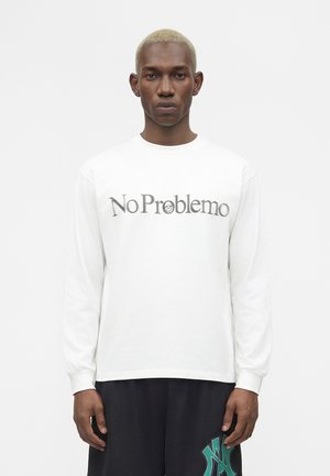 Mand med kort blegnet hår iført en hvid langærmet skjorte med teksten "No Problemo" og sorte bukser med grønt logo.