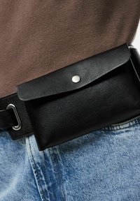 Pochette en cuir noir avec rabat et fermeture à pression argentée, attachée à une ceinture noire. Portée avec un jean denim bleu clair. Surface texturée visible.