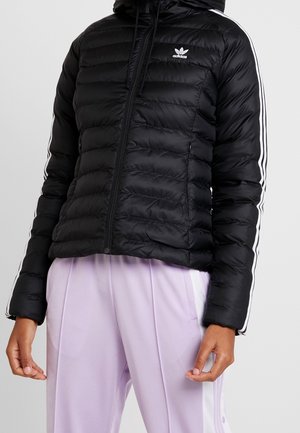 Schwarze Adidas Steppjacke mit weißen Streifen an den Ärmeln, getragen mit hellvioletten Jogginghosen mit seitlichen Knöpfen.