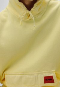 Felpa gialla con cappuccio regolabile, tasca frontale e un piccolo etichetta rossa sulla tasca con la scritta "HUGO" in lettere nere. Tessuto morbido.