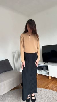 Γυναίκα με μακριά καστανά μαλλιά που φοράει μπεζ μπλούζα με μακριά μανίκια και μαύρη ριγέ μακριά φούστα, στέκεται σε ένα σαλόνι κοντά σε τηλεόραση και καναπέ.