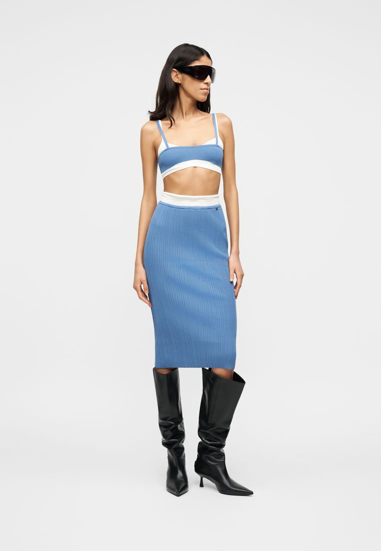 Femme portant un haut court en tricot bleu et une jupe midi assortie avec un bord blanc, des bottes pointues noires montantes jusqu'aux genoux, et de grandes lunettes de soleil noires.