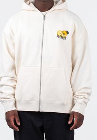 Persona che indossa una felpa con zip color crema, dotata di tasche frontali e un piccolo disegno di un frutto giallo con la scritta "PEGADOR" sul petto sinistro.