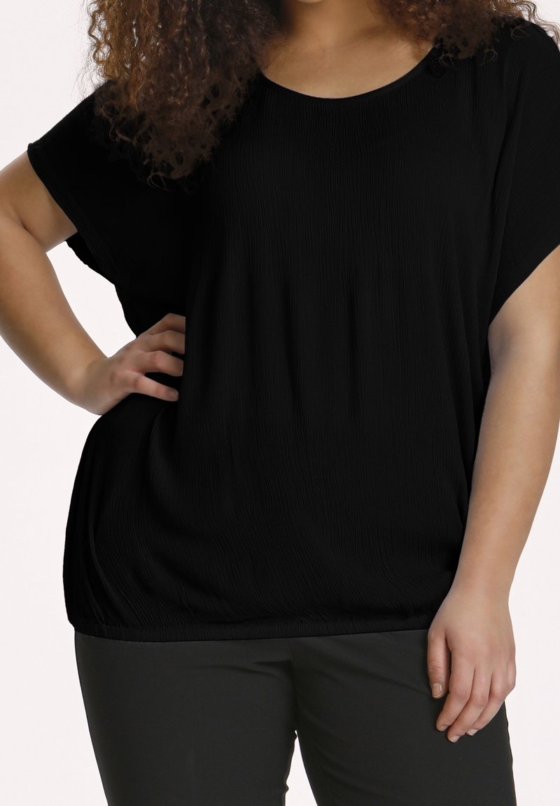 Personne portant un haut noir ample à manches courtes et un pantalon sombre, une main sur la hanche, avec les cheveux bouclés visibles.