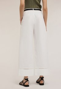 Wide-leg white trousers with a smooth texture, featuring a decorative cutout design at the hems, paired with black strapped sandals.