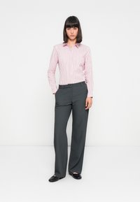 Witte achtergrond; model draagt een roze gestreept overhemd met knopen en grijze wijde broek, met zwarte platte schoenen. Het overhemd heeft een puntige kraag.