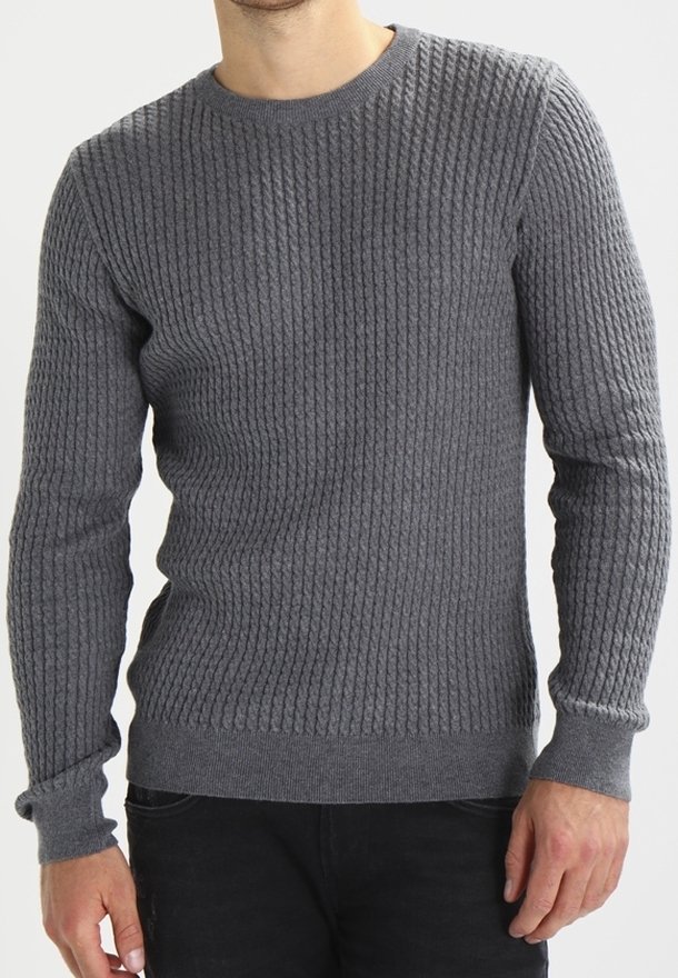 Homme portant un pull gris moulant en maille torsadée et un pantalon noir, debout avec les bras détendus le long du corps sur un fond neutre.