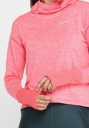 Vrouw draagt een roze sportieve hoodie met lange mouwen en duimgaten en donkere leggings, ze stelt de mouwboord bij tegen een effen achtergrond.