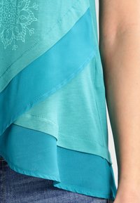 Vrstvený tyrkysový top kombinující matné a průsvitné látky, s květinovým vzorem v horní části a zakřivenými okraji.