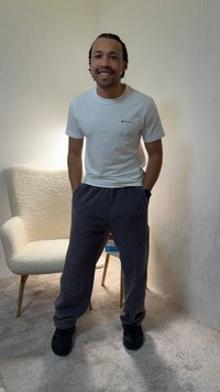Man with braided hair smiles, wearing white t-shirt, gray fleece pants, black shoes, standing in carpeted room near white armchair and small table.