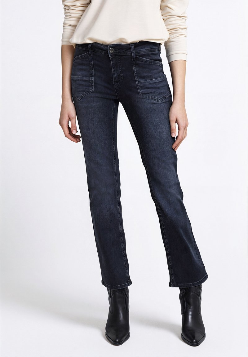 Tmavě modré džíny z denimu s úzkým střihem, vybavené předními kapsami, zapínáním na knoflíky a jemnými detaily šití, doplněné černými kotníkovými botami.