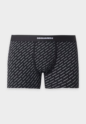 Jeune homme debout pieds nus portant un boxer noir à motifs sur un fond clair uni.