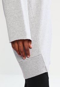 Sudadera gris con una textura suave, diseño holgado y mangas anchas. Los acabados del dobladillo son cortados en bruto. Mano descansando sobre la tela.